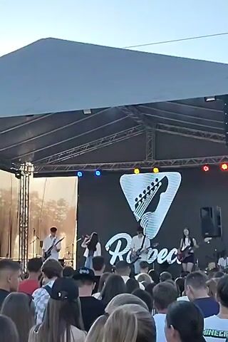 A live band performs on an outdoor stage under a large white canopy. The band consists of at least four young people playing guitars and singing into microphones. A large black screen behind them displays a logo featuring a white guitar silhouette inside a triangle and the word "Respect" in script. A crowd of people is gathered in front of the stage, watching the performance. The sky is visible above the canopy, suggesting it is daytime. The overall scene depicts a concert or music festival.