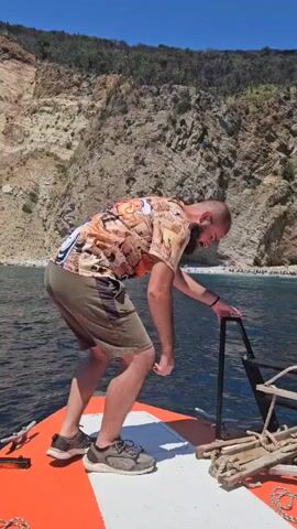 A bald man with a beard wearing a patterned shirt and khaki shorts is crouching on the front of a boat. He is holding onto a metal railing and looking out at a rocky cliff face and a beach with people on it. The water is blue and the sky is clear. There is an anchor and ropes on the boat.