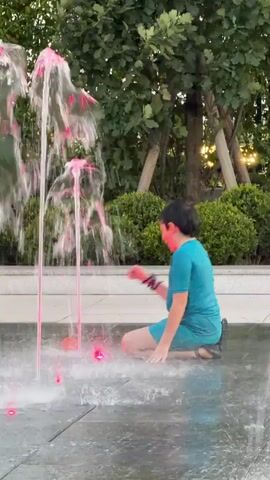 A young boy in a turquoise t-shirt and shorts kneels on a wet, paved surface next to a fountain. Water sprays up from the ground around him, creating a misty atmosphere. He is holding a small, red object in his left hand and appears to be playing with the water jets. The fountain has two metal spouts with water cascading down. Behind the boy, there are green bushes and trees, with sunlight filtering through the leaves. The boy looks at the camera and smiles.