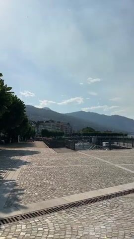 This media file captures a series of scenes along a sun-drenched waterfront. The primary subject is a young woman wearing a straw hat and a green dress, carrying a canvas bag. She is seen smiling and taking a selfie in one shot, indicating a moment of personal enjoyment and perhaps documenting her travels.

The setting appears to be a European lakeside promenade. The ground is paved with cobblestones, forming an attractive pattern. In the background, there are parked cars, trees, buildings, and a prominent body of water. Majestic mountains rise in the distance, suggesting a scenic and possibly mountainous region. The sky is blue with scattered clouds, and the sun is bright, indicating it's daytime, likely late morning or early afternoon, given the quality of light and the shadows.

The activity seems to be a leisurely stroll or exploration of the area. The woman's attire and the overall ambiance suggest a holiday or vacation setting. The presence of a parking lot indicates accessibility for visitors. The waterfront itself features piers or docks, hinting at potential water activities or boat access.

Notable details include the textured cobblestone paving, the lush green trees, the distant mountain range, and the clear blue sky. The woman's cheerful expression conveys a sense of happiness and relaxation. There is no visible text in the media file. The overall impression is one of a beautiful, sunny day in a picturesque Italian lakeside town.