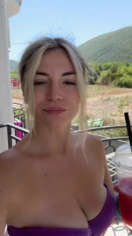 The video opens with a close-up of a young woman with blonde hair, wearing a purple bikini top, holding a plastic cup with a red drink and ice. She is on a balcony. Behind her, a clothes drying rack is visible. The camera pans slightly to the left, revealing a courtyard with a fence, a red wheelbarrow, and a small white building. Then, the view shifts to a swimming pool where two individuals are swimming. One person is splashing water, and another is standing by the pool with a red inflatable ring. The camera then pans back to the woman holding the drink, who is now joined by another young woman wearing a black bikini. They both smile at the camera. The background consists of greenery and hills.