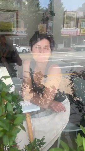A woman with dark hair, wearing a grey tank top and a light-colored skirt, is sitting at a small white table in front of a window. She is leaning on her right hand, with her chin resting on her knuckles, and looking directly at the camera. Her reflection is visible in the window, superimposed over the scene outside. Through the window, a street is visible with buildings, trees, and a white car. To the left of the woman, a man in a grey t-shirt and dark pants is sitting. In the foreground, several potted plants with green leaves are visible. The video appears to be filmed from inside the establishment, looking out.