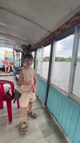 A young boy in a light brown tank top and shorts, wearing sandals, stands on a boat near a window. He holds a red object on a string and plays with it, bringing it towards the camera and then looking out the window. In the background, two adults are seated on the boat, and beyond them, lush green trees and foliage are visible on the shore. The boat interior shows a light blue paneled wall and a silver ceiling with a fan mounted. The lighting suggests it is daytime.