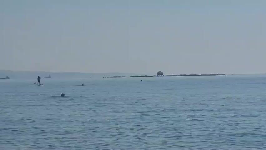 The video shows a wide shot of a calm ocean under a hazy sky. In the foreground, small waves lap at the unseen shore. In the mid-ground, a person stands on a paddleboard, facing away from the camera. To the left of the paddleboarder, a small boat is visible on the horizon. Further to the right, a small island or rocky outcrop is visible with a structure on it, possibly a small building or shed. Several buoys are scattered in the water. The water is a muted blue, and the sky is a pale, uniform gray or light blue. The overall impression is one of a serene, overcast day at sea.