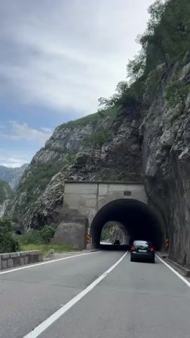 The video shows a car driving through a mountain tunnel on a cloudy day. The tunnel is made of concrete and rock, with lights on the ceiling. Outside the tunnel, the road is paved and has white lines. The surrounding environment is mountainous with green trees. There are other cars on the road. The video appears to be shot from inside a moving vehicle.
