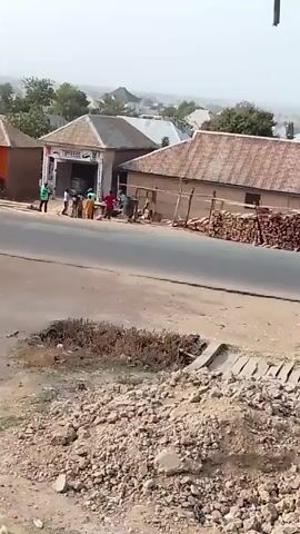 The video shows a street scene in a rural or semi-rural area. There are several buildings on the left side of the road, including a shop with the sign "TOPFEEDS". A group of people are gathered near the shop. Some appear to be carrying bags.  To the right of the shop is a building with wood stacked along its side. The road is paved but appears worn, with potholes and some debris. The foreground shows a patch of rocky, uneven ground. The background shows a distant view of more houses and trees, with a hazy sky above. There is a utility pole visible in the upper right corner.