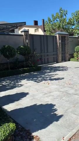 A wide shot of a residential driveway leading to a large, modern gray metal gate with a patterned design. The gate is flanked by brick pillars. To the left of the gate, there is a low gray wall with a dark overhang structure. In front of the wall and gate are two manicured conical topiaries and some rose bushes. The driveway is paved with large, light gray square tiles. The sun casts shadows from trees and foliage onto the driveway. In the background, there are houses with tan walls and brown roofs, and lush green trees. The sky is a clear, bright blue. The video pans slowly to the right, showing more of the gate and the surrounding greenery.