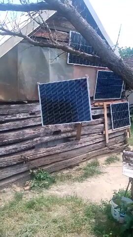 The video shows an outdoor scene with a rustic wooden structure that appears to be a building, possibly a shed or an older house, in Berezynka, Ukraine. The building has a weathered, horizontal plank exterior and a sloped roof. Attached to the exterior and roof are several solar panels, angled to capture sunlight. One large solar panel is prominently displayed in the center, mounted on wooden posts. Two smaller panels are visible higher up on the building, and another is to the right of the large panel. A tree branch extends over the structure, partially obscuring some of the panels. The ground in front of the building is covered with grass and dirt. In the lower right corner, a white bucket with some green plants is visible, suggesting a rural or garden setting. The scene appears to be set during the daytime, with natural light illuminating the area. There are no people visible in the frame. The overall impression is one of a functional, perhaps self-sufficient, setup in a somewhat unrefined, rural environment. The weather appears to be clear or partly cloudy, given the bright sunlight. There is no visible text in the media file.
