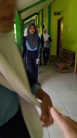 A group of young females wearing hijab and school uniforms walk down a hallway. The hallway has green and yellow walls and a tiled floor. The camera is held by one of the females, and the video shows close-ups of their clothing and faces, as well as the hallway from their perspective. The females are interacting with each other, talking and laughing.