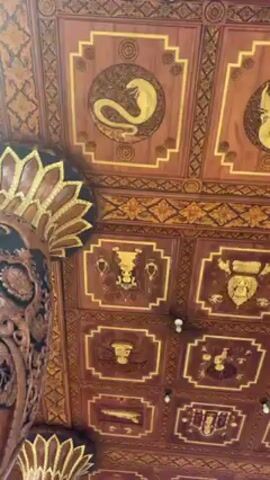 The camera looks up at an ornate, wooden structure with intricate carvings. The ceiling is divided into squares, each containing a golden figure, including dragons and other mythical creatures. The structure is supported by a carved pillar. The background shows a clear blue sky.