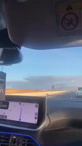 The video, recorded from the interior of a car, shows the dashboard and road ahead. A digital map displaying driving directions is visible. The sunshade is partially lowered, revealing a warning label about airbags. Beyond the windshield is a clear, blue sky with scattered clouds, and a mostly flat, desert-like landscape. Another vehicle, a white van, is visible on the road ahead. An Arabic song plays in the background.