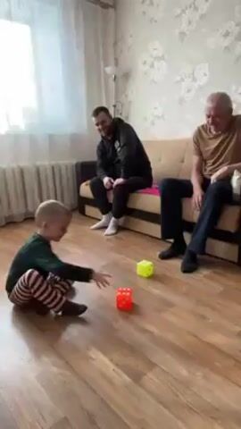 A young boy is playing on the floor of a living room with two dice, an orange one and a yellow one. He wears a dark green shirt and striped brown and white pants. He is facing two men seated on a beige couch. One man is young, wearing a black hoodie, and the other man is elderly, wearing a tan shirt and dark slacks. The boy crawls toward the dice, picks up the orange one, and sits down while manipulating the cube. The young man watches the boy and smiles. The older man also smiles and watches the boy. The room is decorated with floral wallpaper and has a window with white blinds. A floor lamp stands in the corner. The floor is made of light-colored wood.