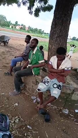The video shows a group of young men outdoors in a somewhat rural setting. In the foreground, three men are sitting on a concrete ledge under a large tree. One man is wearing a green t-shirt, another a patterned shirt, and the third a white shirt with a collar and shorts. The ground is dirt and littered with debris. In the background, a few other people are visible, some near a rudimentary structure made of corrugated metal and wood. One person is standing on what appears to be the roof or upper part of the structure. The atmosphere seems casual, with the men appearing to be resting or socializing. The overall impression is of a warm, possibly humid day in a developing area.