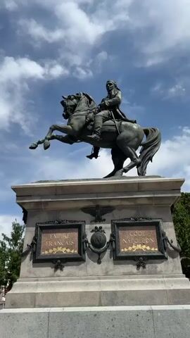 The video shows a low-angle, close-up, full-shot of a large bronze equestrian statue of a man, likely Napoleon Bonaparte given the inscription on the base. The statue is mounted on a tall, grey stone pedestal. The pedestal has two framed inscriptions, one on the left that reads "EMPIRE FRANCAIS" and one on the right that reads "CODE NAPOLEON". There are also decorative elements on the pedestal, including what appears to be a laurel wreath and an eagle. The statue is detailed, with the man dressed in military attire and his horse captured in mid-stride, with its front legs raised. The sky behind the statue is blue with scattered white clouds. Trees can be seen on the right side of the frame. The lighting suggests it is daytime, possibly late morning or early afternoon.
