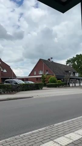 A short vertical video shows a static outdoor view. In the foreground, there's a gray asphalt road and a section of a sidewalk made of light-colored pavers. A dark green bush hedge separates the road from a parking area where parts of two dark-colored cars are visible on the left. Beyond the parking area, there's a two-story brick building with a dark tiled roof. It has white-framed windows and a chimney visible on the roof. To the right of this building, a smaller, darker structure with large windows and an awning is attached, possibly a restaurant or shop. The background features more greenery, including trees on the far right. The sky is partially cloudy with patches of blue visible. Throughout the video, a loud, continuous, high-pitched ringing sound is heard, resembling an alarm or bell.