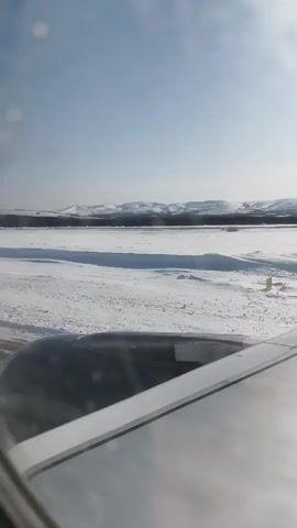 The video, shot from an airplane window, shows a vast expanse of snowy landscape under a bright sky.  Mountains are visible in the distance. The wing of the plane is partly visible in the foreground. The environment appears cold and icy. There are no people visible in the video. The plane appears to be moving or about to take off.