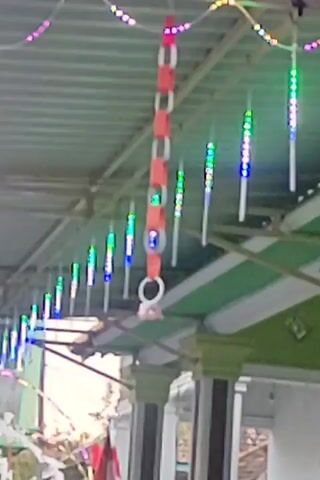 The video shows a close-up, low-angle shot of an outdoor covered area. The roof is corrugated metal. Hanging from the roof are several strands of decorative lights. Most of these lights are small, cylindrical, and emit a changing sequence of colors, appearing to be LED lights. Some are white or translucent tubes with colored lights inside. One strand is a chain of red and white paper rings. There are also a few larger, flat, colorful lights strung across the top. Below the roof structure, there are visible architectural elements, including green and white trim, and what appear to be pillars. The background is slightly out of focus, but suggests more structures and some foliage. The lighting suggests it might be dusk or a dimly lit area. No people are visible. The overall impression is of a decorated outdoor space, possibly for a celebration.