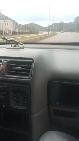The video shows the interior of a car as it drives on a road. The dashboard is visible, including the steering wheel, radio, air vents, and a clock. A hand is seen on the steering wheel. The view outside the car shows buildings, trees, and a road with other vehicles. The lighting suggests it is daytime.