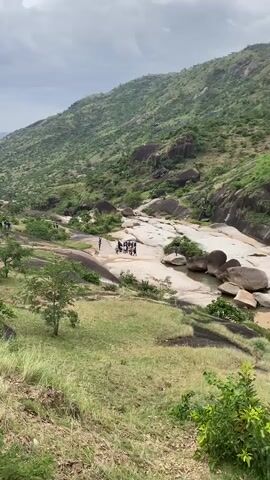 The video presents a scenic landscape with a lush, green hillside in the background under an overcast sky. The foreground features a rocky, pale-colored flat area dotted with pools of water and interspersed with green vegetation. A group of approximately 15 people, mostly dressed in casual clothing, are gathered on the flat rocky surface. Some individuals are walking around while others appear to be standing and talking. The camera angle is from a higher vantage point, looking down upon the group and the rocky terrain. The setting suggests a natural environment, possibly a park or a hiking trail, and the group appears to be exploring or socializing in the area.