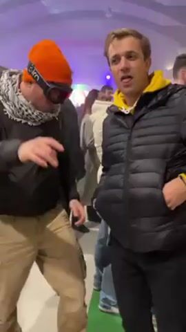 Two young men are dancing at what appears to be an indoor music festival or party. The man on the left wears an orange ski hat and goggles, a black jacket, a scarf, and khaki pants. He sticks his tongue out. The man on the right wears a black puffer jacket with a yellow hoodie underneath, and black pants. Both men make hand gestures. The scene is brightly lit with colored lights. Other people are visible in the background. The floor is a light color.