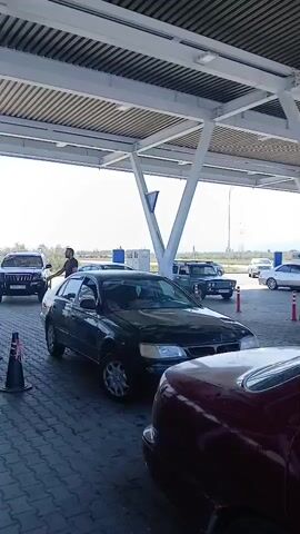 The video shows a line of cars at what appears to be a gas station or car wash, under a covered structure. In the foreground, a dark-colored sedan is parked, with its rear partially visible. Next in line is a dark green sedan, also parked. Further back, a black SUV is visible, and beyond that, more vehicles are present. A person, appearing to be a male wearing shorts and a t-shirt, is walking away from the SUV towards the left of the frame, past some orange and black traffic cones. The ground is paved with interlocking bricks. Green containers, possibly for fuel or other liquids, are stacked to the left of the frame. The sky is bright and clear, suggesting it is daytime.
