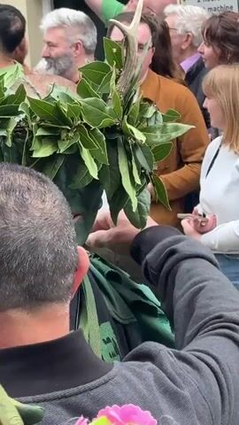 A man with his face and hands painted green, wearing a leafy garment and a laurel wreath with antlers on his head, hugs another man in a crowd. The man in green holds a stick and a flower is tucked into his garment. Spectators surround them, some holding drinks and phones. The scene takes place outside a building with a sign that says "Throught."
