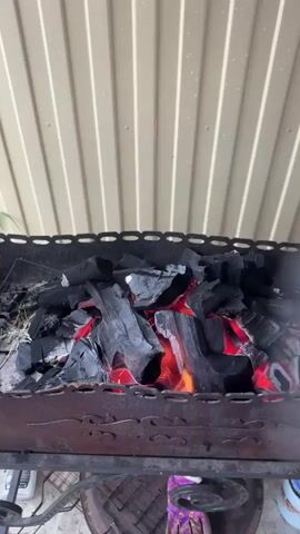 The video shows a close-up, static shot of glowing charcoal in a grill. The charcoal is piled high and appears to be actively burning, with red and orange embers visible underneath the black exterior. The grill itself is made of dark metal with decorative patterns on the sides. In the background, a cream-colored corrugated metal surface forms the ceiling or wall of an outdoor area. There are no people or animals visible in the frame, and no specific actions or interactions are taking place beyond the natural burning of the charcoal. Some sparks can be seen rising from the coals. A blurred object, possibly a hand with a glove, briefly moves across the right side of the frame at the beginning of the video, creating a large plume of smoke.
