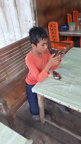 A young boy wearing an orange long-sleeved shirt and blue pants kneels at a table in an outdoor setting. He is holding a smartphone in his hands and appears to be looking at it intently. On the table in front of him, there are some pieces of food, possibly fruit or nuts, and a slice of what looks like banana. The table has a light green, textured surface. Behind the boy, there is a wooden bench with vertical slats. In the background, several orange plastic chairs are visible, and there are containers on a shelf. The environment appears to be a simple outdoor dining area, possibly a food stall or open-air restaurant.