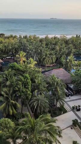 The video is an aerial shot of a tropical resort. In the foreground, there are palm trees and a paved area with a water feature. In the midground, there are more palm trees, buildings with thatched roofs, and a swimming pool with lounge chairs. In the background, there is a sandy beach, the ocean, and a distant island. The sky is a soft orange and yellow, indicating either sunrise or sunset. Several people can be seen walking around the resort grounds.