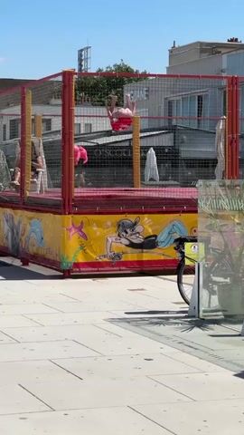 A group of young people are on a trampoline with a red surface and a yellow safety net. The trampoline is decorated with cartoonish images of sea creatures and a person. In the background, there are buildings, trees, and a clear blue sky. A bicycle is parked to the right of the trampoline. One person in a red shirt is in the center of the trampoline, facing away from the camera and appearing to be sitting or kneeling. Another person in a pink shirt is in the background on the left side of the trampoline, also appearing to be sitting. The scene is illuminated by bright sunlight, suggesting a daytime setting.