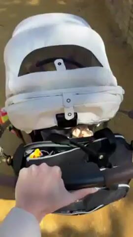 The video shows a first-person view of someone pushing a white and black baby stroller on a dirt path. The stroller has a white hood with a mesh window. A black diaper bag hangs from the stroller's handle. The ground is covered in light brown dirt and some shadows. The video is stable and the focus is on the stroller. The baby inside makes cooing sounds.