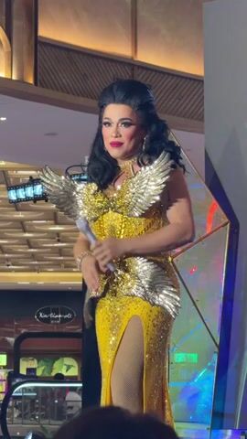 A drag performer stands on a stage, singing into a microphone. They are wearing a sparkly yellow gown with a high slit, embellished with gold wings on the shoulders and waist. The performer has dramatic makeup and dark, voluminous hair.

The setting appears to be an indoor event, possibly a mall or entertainment venue, given the presence of slot machines visible in the background. The stage has a geometric backdrop with iridescent panels and lighting. The performer's expression is animated and engaging as they sing. The overall mood is celebratory and performative.

Visible text includes "Xin" on a sign in the background. The time of day is not discernible from the image. The location is specified as Cebu City, Philippines.