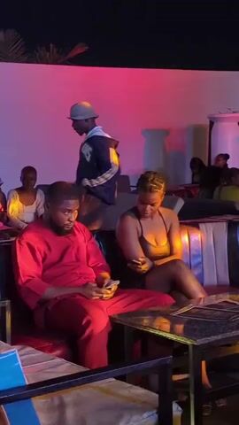 This media file depicts a dimly lit outdoor seating area at Bar Jirgi Summa, Nigeria. The primary subjects are a Black man and a Black woman seated on a cushioned bench. The man, positioned on the left, wears a red garment and is looking down at his phone. The woman, on the right, wears a black top and also appears to be focused on her phone. Behind them, other patrons are visible in the background, seated at tables and on similar benches.

The scene is illuminated by changing colored lights, primarily shades of blue, green, and purple, suggesting nighttime. The overall ambiance appears relaxed and casual. The presence of tables and seating indicates a social gathering or a place for leisure. The background features architectural elements and some greenery, with a palm frond visible on the far left, contributing to an outdoor setting.

There is no visible text in the media file. The time of day is clearly night, and the weather is not discernible. The emotions of the subjects are not explicitly conveyed due to their focus on their phones and the dim lighting. The location cues, such as the palm frond and the name of the establishment, strongly suggest Nigeria as the setting.