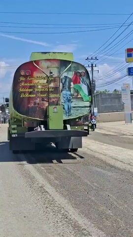 A rear view of a green water tanker truck driving on a dusty road. The truck has a large graphic on its back, depicting what appears to be a scene of conflict on the left, with an image of a person holding a Palestinian flag on the right. The graphic also contains yellow script in a foreign language. A motorcycle with two riders is visible in the background. The sky is blue and clear.