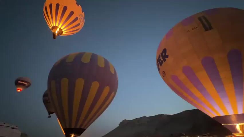 The video shows multiple hot air balloons in the sky at dusk. The balloons are lit from within, illuminating their colorful patterns against the darkening blue sky. Some of the balloons are close to the camera, showing detailed designs of purple and yellow stripes, and orange and pink stripes with the word "BUTTERFLY" visible on one. In the background, the silhouettes of hills or mountains are visible, along with a few distant lights and vehicles on the ground. The scene conveys a sense of peaceful wonder and anticipation.