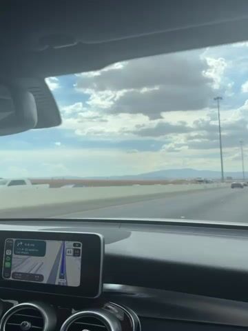 This video shows a first-person view from a car driving on a highway. The car's dashboard is visible in the lower left corner, with a navigation screen displaying a map and route. The road ahead is multi-lane with other vehicles, including a car carrier truck, several cars, and some trucks. A concrete barrier separates the lanes of traffic and extends into the distance. Beyond the barrier, a flat, arid landscape with distant mountains under a cloudy sky is visible. A speed limit sign indicating 65 mph is seen on the right side of the road. Green highway signs with route information are also present. The overall environment appears to be a highway in a desert or semi-arid region.