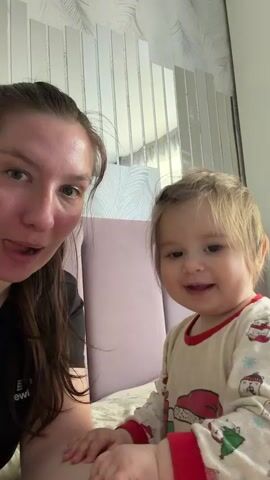 A woman and a young child are seated on a bed. The woman is on the left side of the frame, wearing a black shirt, with her dark hair pulled back. The child is seated on the right side of the frame, wearing pajamas with Christmas-themed images. The child's hair is light blonde. The woman smiles at the camera. The child looks at the camera with a serious expression. The headboard is light purple. Behind them, the wall is decorated with vertical silver panels.