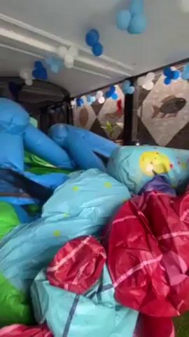 The video shows an inflatable bouncy house placed under a covered patio, decorated with blue and white balloons. The bouncy house, which appears deflated, is made up of various blue, red, and green sections. A tile wall with a fish design is visible in the background. Loud music is playing.