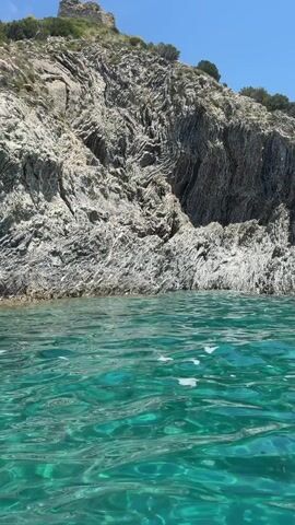 The camera shows a view from the water towards a rocky cliff and hillside. The water is clear and turquoise, with gentle ripples. The cliff is composed of light gray rock with darker gray striations, creating a folded or layered appearance. Some sparse greenery and trees are visible on top of the cliff, and a small stone structure that looks like a ruined building can be seen at the peak of the hillside. The sky is blue and clear.