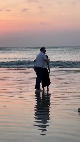 Two people are dancing on a beach at sunset. The man is wearing a white shirt and dark pants, and the woman is wearing a white top and a dark skirt. They are reflected in the wet sand. The sky is a soft pink and orange, and the ocean is calm with gentle waves.