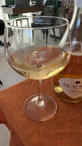The video shows a partially filled wine glass and a bottle of Ca' Di Rajo Prosecco D.O.C. Treviso Rosé on an orange tablecloth. The wine in the glass appears light in color. The wine bottle is next to the glass. In the background, there is a desk with a laptop and a green chair. A black backpack is on the floor. Someone is speaking Russian in the background.