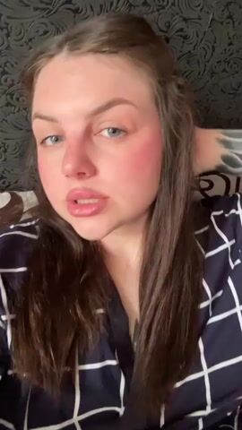 A woman is lying down, possibly in bed, wearing a blue and white patterned robe. She has long brown hair, blue eyes, and a nose ring. She is looking at the camera, smiling, and touching her hair. In the background is a dark patterned headboard.
