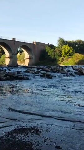 A video shows a wide river with a rocky bottom and small rapids. On the left side of the screen, a stone arch bridge spans the river, with green trees and foliage visible behind it. The water is a deep blue, and the sky is clear and bright. The camera pans across the river, capturing the movement of the water and the natural scenery.