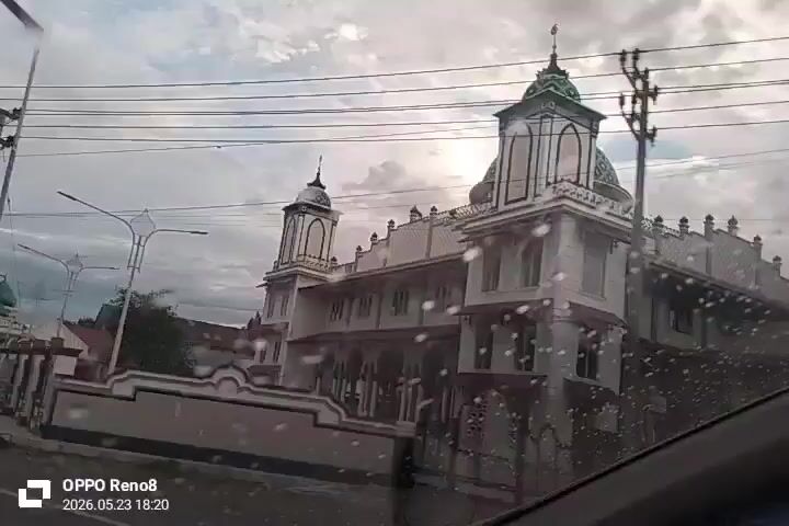 The video appears to be shot from inside a moving vehicle, possibly a car. The footage shows a view of a street in a town or city on what seems to be a cloudy day. The vehicle drives past buildings, some of which appear to be shops, and utility poles with electrical wires. A mosque with a distinctive green dome and minarets is visible. Other vehicles, including cars and motorcycles, are also present on the road. Water droplets on the windshield suggest it might have been raining or that the window is wet. A timestamp indicates the video was taken in May 2026.