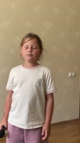 In this video, a young girl with blonde hair is standing in front of a beige, floral-patterned wallpaper. She is wearing a white t-shirt with pink trim and pink shorts. She is holding a black microphone in her right hand and appears to be singing. Her facial expression suggests she is engaged in the activity, possibly with a mix of concentration and enjoyment. Behind her, a light switch is visible on the wall. The scene suggests an indoor setting, possibly a home. The lighting is neutral, not indicating a specific time of day. The overall mood appears to be lighthearted and playful, centered around the child's performance. There is no visible text in the frame.