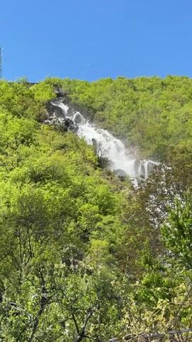 The video shows a waterfall cascading down a rocky hillside surrounded by lush green trees under a clear blue sky. The water is white and foamy as it flows over the rocks. At the top of the waterfall, there is a tunnel-like structure visible. The camera zooms in slightly on the waterfall, providing a closer view of the water and the surrounding vegetation.