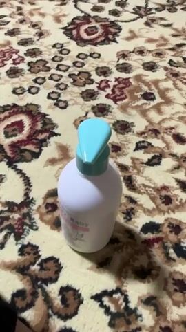 A person's hand is shown in the foreground, blurred and moving. In the mid-ground, a white plastic bottle with a turquoise pump dispenser is upright. The bottle has a label with faint text and a drawing. The background is a close-up of a patterned rug with intricate designs in shades of beige, red, and black. The overall scene appears to be indoors, likely a home environment, given the presence of the rug. The lighting suggests it could be daytime. The activity is the dispensing of liquid from the bottle into the person's hand, indicated by a drop of clear liquid falling from the dispenser. The focus is on the action of using the product.
