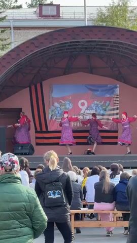 The video shows a dance performance by six girls on a stage. They are wearing matching purple dresses with white aprons and red headscarves. They dance in unison to music being played, in front of a banner in Russian that includes the date “9 May 1941-1945.” The stage is covered by a large roof and is set outside with a crowd of spectators seated in front of it, watching the performance.