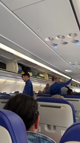 The video shows the interior of an airplane cabin. Passengers are seated in blue seats, and two flight attendants in blue shirts are visible. One flight attendant is standing near the front of the cabin with his arms folded, while the other is reaching up to adjust luggage in an overhead bin. The bins are partially filled with various bags and suitcases. The cabin lighting is bright, and the ceiling panels and air vents are visible.