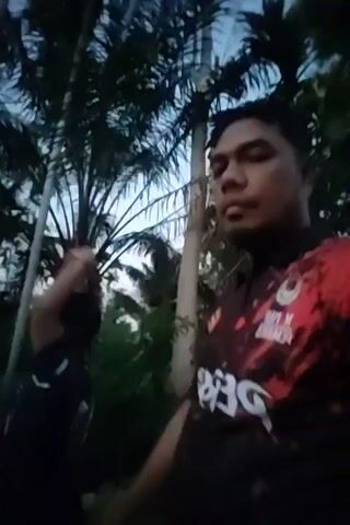 Two people are visible in the video. A man, presumably the videographer, is in the foreground, wearing a red and black patterned shirt. To his left and slightly behind him, is a child in a blue shirt. Both are looking to the left, away from the camera. They are surrounded by tall trees and greenery, possibly a park or garden setting. The sky in the background is light blue, suggesting it may be early evening or late afternoon. The man is seen raising his hand to his mouth, possibly to smoke or eat something. The child has their hand to their mouth, possibly chewing or thinking.